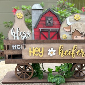 Puede incluir: Carro de madera con tem&aacute;tica de granja con elementos decorativos. Cuenta con un granero rojo, una vaca y un letrero que dice "Hey Heifer". El carro tiene ruedas de madera y est&aacute; decorado con flores.