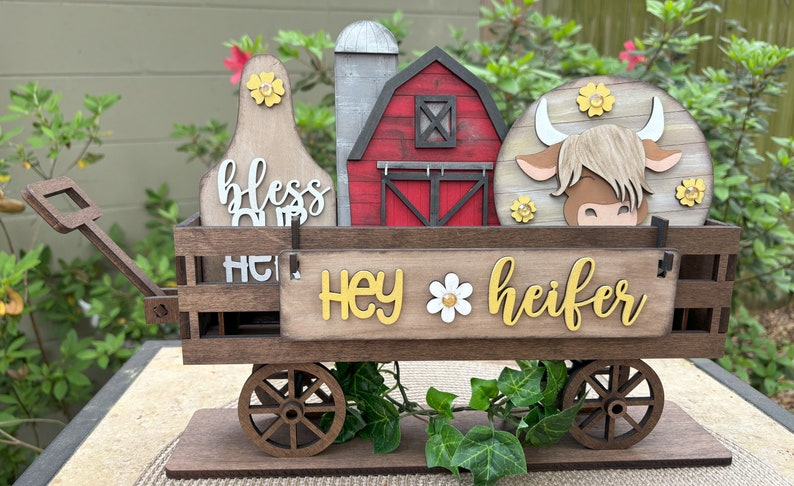 Puede incluir: Carro de madera con tem&aacute;tica de granja con elementos decorativos. Cuenta con un granero rojo, una vaca y un letrero que dice "Hey Heifer". El carro tiene ruedas de madera y est&aacute; decorado con flores.