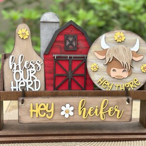 Puede incluir: Decoraci&oacute;n de madera con tem&aacute;tica de granja con un granero, un silo y una cabeza de vaca. El letrero dice "bendice a nuestro reba&ntilde;o" y "Hey heifer". La cabeza de la vaca tiene el texto "Hey there". Se incluyen acentos de flores amarillas.