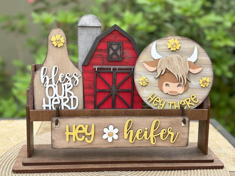 Puede incluir: Decoraci&oacute;n de madera con tem&aacute;tica de granja con un granero, un silo y una cabeza de vaca. El letrero dice "bendice a nuestro reba&ntilde;o" y "Hey heifer". La cabeza de la vaca tiene el texto "Hey there". Se incluyen acentos de flores amarillas.