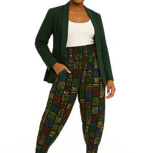 May include: A woman wearing a dark green blazer, white top, and patterned pants. The pants feature a repeating geometric design in red, yellow, blue, and green on a black background. She is also wearing a matching head wrap and black high-heeled sandals.