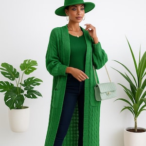 Könnte beinhalten: Eine Frau in einem grünen Outfit, bestehend aus einem langen Zopfstrick-Cardigan, einem Oberteil und einem breitkrempigen Hut. Sie trägt auch dunkelblaue Jeans, Goldschmuck und High Heels. Eine kleine grüne Handtasche mit goldener Kette ist ebenfalls zu sehen.