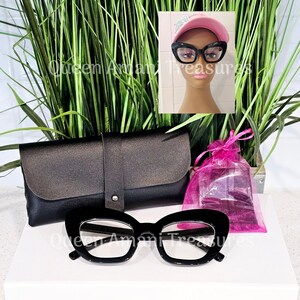 May include: Black cat eye glasses with a clear lens in a black case and a pink drawstring bag. The glasses are on a white surface.
