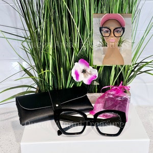 May include: Black cat-eye glasses with a gold accent on the bridge of the frame. The glasses are on a white surface next to a black leather glasses case and a pink mesh bag.