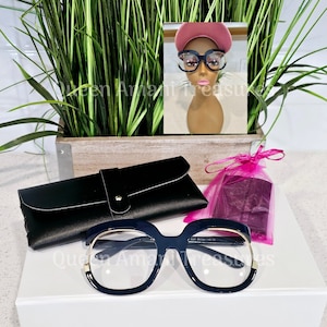 May include: A set of black oversized glasses with gold accents, a black glasses case, and a pink pouch. The glasses are on a white box with the text "Queen Amani Treasures" on the front.