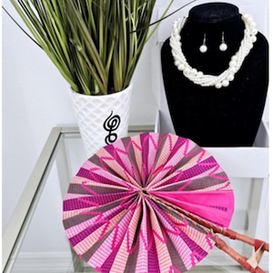 May include: A pink and brown hand fan with a wooden handle. The fan is folded and has a geometric pattern. The fan is on a glass table with a white vase and green plants in the background.