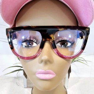 May include: A mannequin wearing a pink baseball cap and tortoise shell glasses with pink tinted lenses. The mannequin is wearing a silver choker necklace with a silver pendant shaped like the continent of Africa.