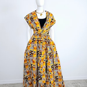 Könnte beinhalten: Ein lebendiges, gemustertes Kleid an einer Schaufensterpuppe. Das Kleid hat ein gelbes, schwarzes und weißes geometrisches Design. Es hat ein Oberteil im Wickelstil und einen vollen, fließenden Rock. Eine goldfarbene Halskette mit Anhänger ist sichtbar.