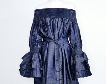 Puede incluir: Vestido azul oscuro con hombros descubiertos, escote fruncido y mangas largas con volantes en capas. El vestido presenta un cinturón a juego y se exhibe en un maniquí. El vestido está hecho de una tela similar al denim.