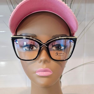 May include: Black cat-eye glasses with gold accents and blue light blocking lenses. The glasses are on a mannequin head wearing a pink baseball cap.