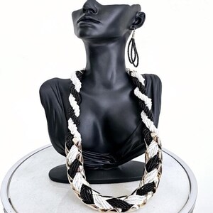May include: A black and white beaded necklace with a gold chain accent. The necklace is twisted and has a geometric pattern. The necklace is displayed on a black mannequin bust.