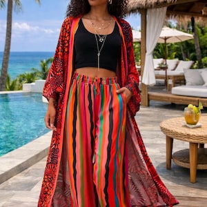 Op de afbeelding: Een vrouw draagt een zwarte crop top, een kleurrijke gestreepte broek en een lange rode kimono met patroon. De broek heeft verticale strepen in verschillende kleuren. De achtergrond toont een zwembad, palmbomen en een tropische omgeving.