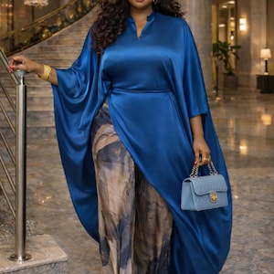May include: A woman in a royal blue satin dress with a cape-like design and a matching blue handbag. The dress is paired with patterned pants in shades of brown and gray. Gold bracelets and earrings complete the look.