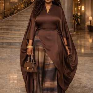 May include: A brown satin wrap dress with wide-leg pants featuring a watercolor-like pattern. The outfit includes a matching brown handbag and gold bracelets. The background shows a grand staircase and a chandelier.