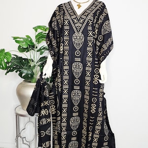 Puede incluir: Un vestido caftán negro y dorado estampado con un bolso con cordón a juego. El vestido presenta un cuello en V y un diseño repetido de máscaras tribales y formas geométricas. El vestido se exhibe en un maniquí.