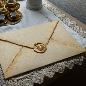 Può includere: Una busta in stile vintage con un bordo strappato e un sigillo di cera dorata con un design a corona d'alloro. La busta è di colore beige chiaro, con una superficie strutturata. Il sigillo è circolare e aggiunge un tocco di eleganza.
