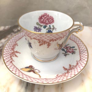 Könnte beinhalten: Ein weißes Tee-Set mit Blumendesign. Die Teetasse hat eine rosa Blume und goldene Verzierungen. Die Untertasse hat ein rosa Gittermuster und eine Schmetterlingsillustration. Ein klassisches Teeservice.