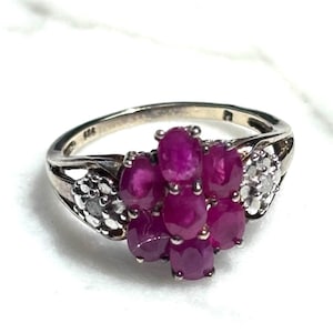 Op de afbeelding: Een zilverkleurige ring met een cluster van zeven ovaal geslepen dieproze robijnen in een bloemmotief, geflankeerd aan weerszijden door een kleine cirkelvormige groep van piepkleine heldere witte steentjes in de ringband met een keurmerk.
