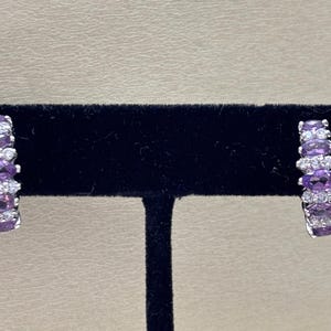 Puede incluir: Un par de pendientes de aro plateados. Cada pendiente presenta una fila de piedras preciosas moradas de corte ovalado alternando con piedras blancas redondas más pequeñas. Los pendientes se exhiben sobre una superficie de terciopelo negro.