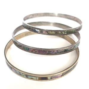 May include: Three thin metal bangle bracelets stacked together. Each bracelet features a smooth silver-toned metal frame inlaid with a continuous band of iridescent abalone shell segments displaying shades of green, purple, and blue throughout.