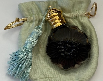Puede incluir: Una pequeña botella de vidrio de color morado oscuro con una tapa dorada y un diseño en forma de flor. La botella está en una bolsa de terciopelo verde claro con un borla azul.