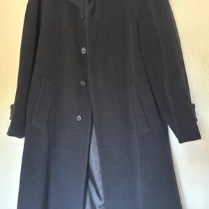 May include: A long, black overcoat with a classic design. The coat features a notched collar, button closure, and front pockets. The sleeves have buttoned cuffs. The coat is hanging on a hanger.