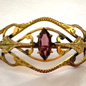 May include: A gold-toned brooch with an intricate design featuring a purple gemstone in the center.