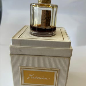 Op de afbeelding: Een klein, doorzichtig glazen flesje parfum met een gouden dop en een wit etiket met de tekst "Jasmin Le Galion 1/2 FL OZ". Het flesje staat op een witte doos met een geel etiket waarop "Jasmin Le Galion 1/2 FL OZ" staat.