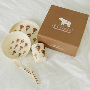 May include: A brown box with the text "LU & KEN CO. Kids bamboo dinnerware" and a white bear illustration. The box contains a set of bamboo dinnerware with a hot air balloon design. The set includes a bowl, a plate, a cup, a spoon, and a fork.