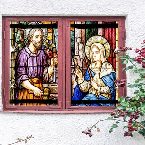 以下が含まれることがあります： 男性と女性を描いたステンドグラスの窓。男性は紫色のローブを着て、ハンマーを持っています。女性は青いローブを着て、糸巻きを持っています。窓は白い壁に設置されており、手前にピンクのバラの茂みが植えられています。