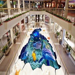 May include: A 3D floor decal depicting an underwater scene with a shark, dolphins, and coral. The decal is placed on a floor in a shopping mall.