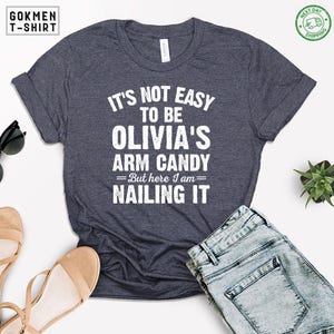 May include: Dark gray t-shirt with the text "IT'S NOT EASY TO BE OLIVIA'S ARM CANDY =But here I am= NAILING IT" in white. The shirt is paired with blue jeans and sandals. The shirt has rolled-up sleeves.