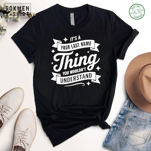 May include: Black t-shirt with the text "IT'S A YOUR LAST NAME Thing YOU WOULDN'T UNDERSTAND" in white. The shirt has a crew neck and short sleeves. The image also shows a pair of white shoes, a brown hat, and blue jeans.