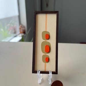 Pode incluir: Uma impressão de arte abstrata emoldurada com uma moldura marrom e uma tela branca com três formas laranja e verde e duas listras laranja.
