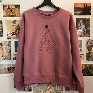 May include: Mauve-colored sweatshirt with a crew neck and long sleeves. The front features a minimalist line drawing of a person carrying a child. The sweatshirt is hanging on a black hanger against a wall decorated with various art prints.
