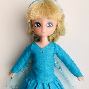 K&ouml;nnte beinhalten: Eine blaue und wei&szlig;e Puppe mit blonden Haaren, die ein blaues Kleid und ein blaues Schleifenband tr&auml;gt.