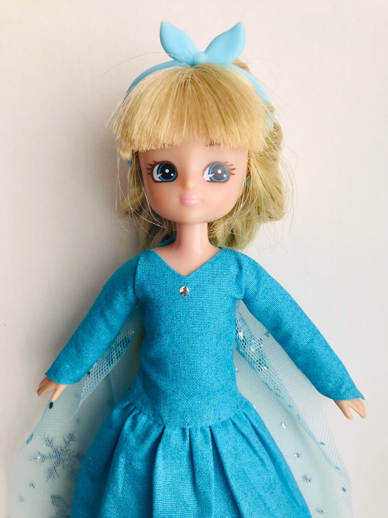 K&ouml;nnte beinhalten: Eine blaue und wei&szlig;e Puppe mit blonden Haaren, die ein blaues Kleid und ein blaues Schleifenband tr&auml;gt.