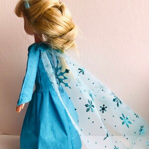 K&ouml;nnte beinhalten: Ein blaues Kleid mit einem langen, flie&szlig;enden T&uuml;llcape mit Schneeflockenmuster. Die Puppe hat blondes Haar, das zu einem Zopf geflochten ist, und eine blaue Haarklammer.