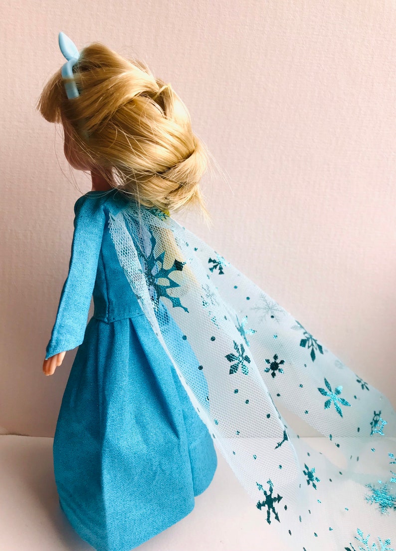 K&ouml;nnte beinhalten: Ein blaues Kleid mit einem langen, flie&szlig;enden T&uuml;llcape mit Schneeflockenmuster. Die Puppe hat blondes Haar, das zu einem Zopf geflochten ist, und eine blaue Haarklammer.