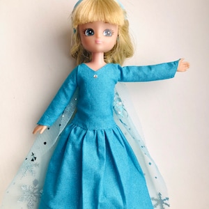 K&ouml;nnte beinhalten: Eine blaue Puppe, die ein blaues Kleid mit einem T&uuml;llcape und einer blauen Schleife im Haar tr&auml;gt. Das Cape hat ein Schneeflockenmuster.