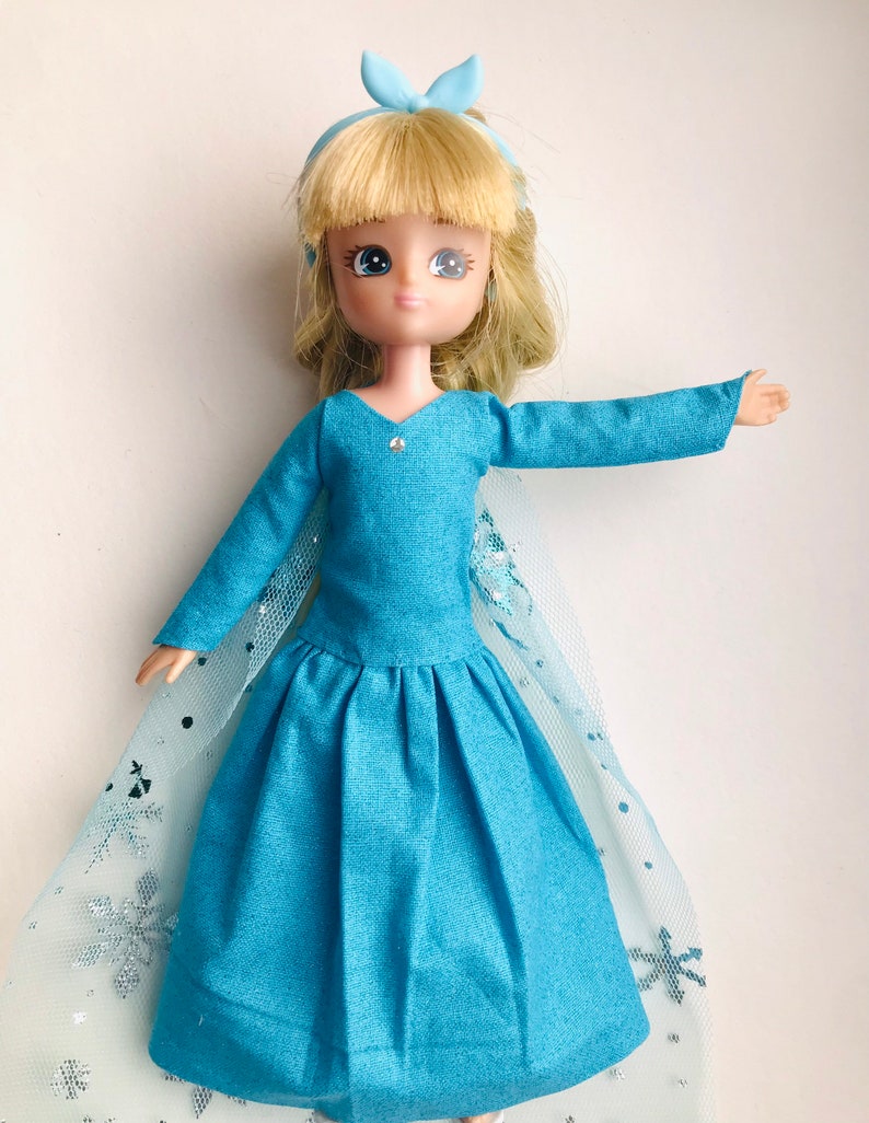 K&ouml;nnte beinhalten: Eine blaue Puppe, die ein blaues Kleid mit einem T&uuml;llcape und einer blauen Schleife im Haar tr&auml;gt. Das Cape hat ein Schneeflockenmuster.