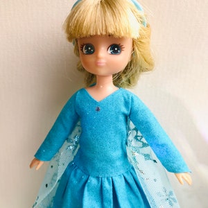 K&ouml;nnte beinhalten: Eine blaue und wei&szlig;e Puppe, die ein blaues Kleid mit einer wei&szlig;en Spitzen&uuml;berlage und einer blauen Schleife im Haar tr&auml;gt.