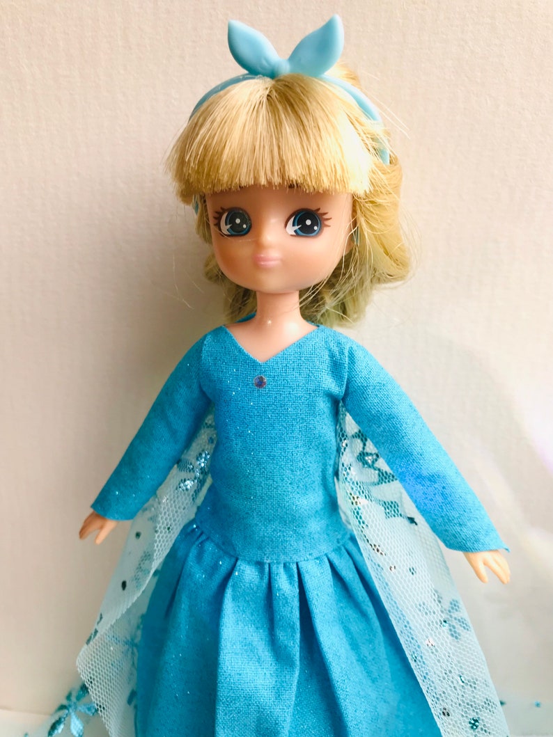K&ouml;nnte beinhalten: Eine blaue und wei&szlig;e Puppe, die ein blaues Kleid mit einer wei&szlig;en Spitzen&uuml;berlage und einer blauen Schleife im Haar tr&auml;gt.