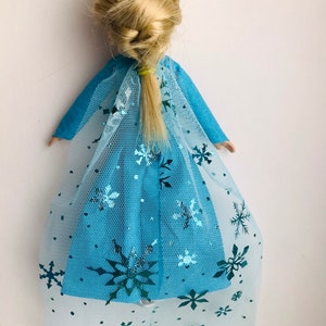K&ouml;nnte beinhalten: Eine blaue Puppe mit einem langen blonden Zopf, die ein blaues Kleid mit einem T&uuml;llcape tr&auml;gt, das mit silbernen Schneeflocken verziert ist.