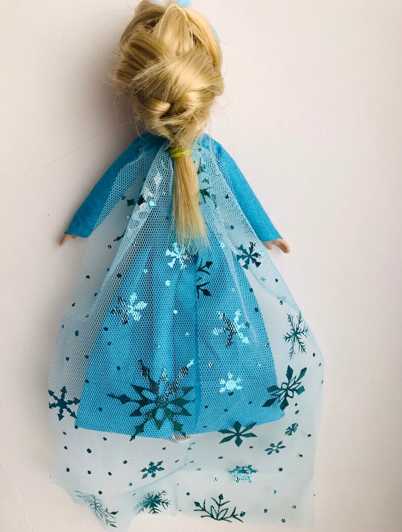 K&ouml;nnte beinhalten: Eine blaue Puppe mit einem langen blonden Zopf, die ein blaues Kleid mit einem T&uuml;llcape tr&auml;gt, das mit silbernen Schneeflocken verziert ist.