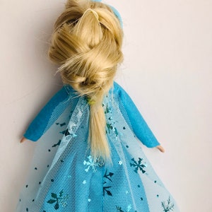 K&ouml;nnte beinhalten: Eine blaue und wei&szlig;e Puppe mit einem blonden Zopf und einer blauen Schleife im Haar. Die Puppe tr&auml;gt ein blaues Kleid mit einem wei&szlig;en T&uuml;ll&uuml;berzug und Schneeflockenmustern.