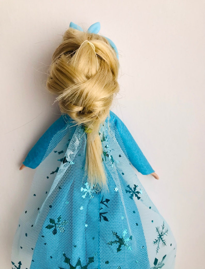 K&ouml;nnte beinhalten: Eine blaue und wei&szlig;e Puppe mit einem blonden Zopf und einer blauen Schleife im Haar. Die Puppe tr&auml;gt ein blaues Kleid mit einem wei&szlig;en T&uuml;ll&uuml;berzug und Schneeflockenmustern.