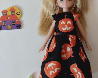 Puede incluir: Una muñeca con cabello rubio que lleva un vestido negro con calabazas naranjas. Recortes decorativos de un murciélago, una casa embrujada y una calabaza están junto a la muñeca. La muñeca lleva zapatos dorados.
