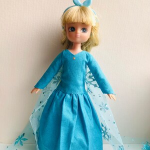 K&ouml;nnte beinhalten: Eine blaue Puppe, die ein blaues Kleid mit einem Spitzenumhang und einer blauen Schleife im Haar tr&auml;gt. Die Puppe hat blonde Haare und blaue Augen.