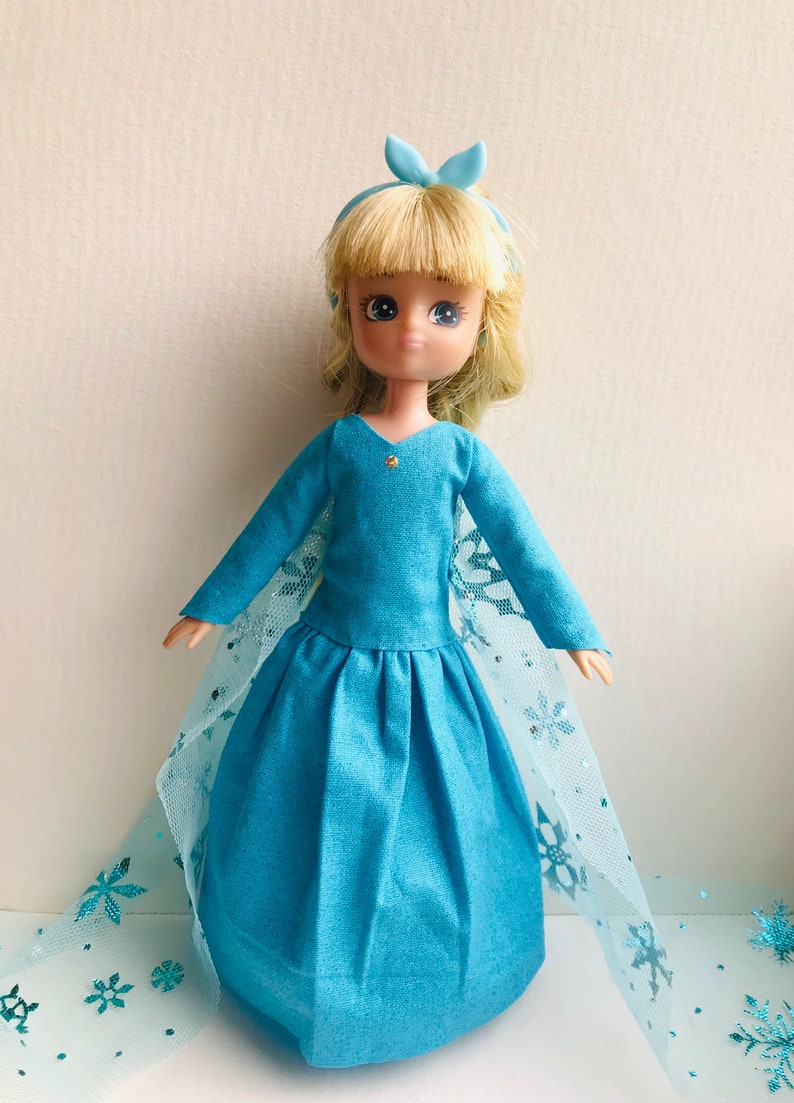 K&ouml;nnte beinhalten: Eine blaue Puppe, die ein blaues Kleid mit einem Spitzenumhang und einer blauen Schleife im Haar tr&auml;gt. Die Puppe hat blonde Haare und blaue Augen.